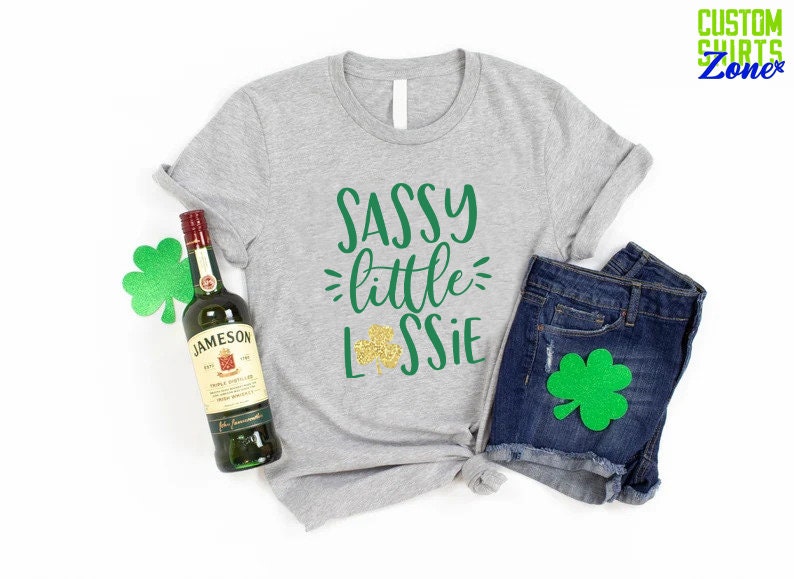 K&ouml;nnte beinhalten: Ein graues T-Shirt mit dem Text "Sassy little Lassie" in Gr&uuml;n und goldenen Glitzer. Das Shirt wird mit Jeans-Shorts und einer Flasche Jameson Irish Whiskey kombiniert. Es gibt gr&uuml;ne Kleebl&auml;tter, die um das Bild herum verstreut sind.