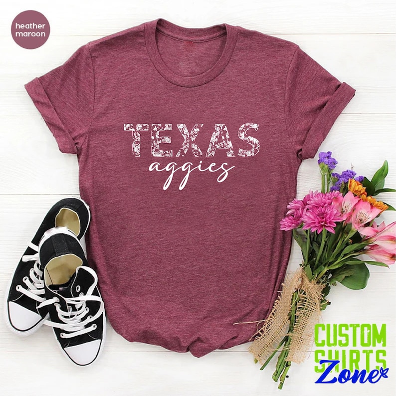 Może przedstawiać: Koszulka w kolorze brązowo-czerwonym z białym wzorem kwiatowym, na kt&oacute;rym widnieje napis "TEXAS aggie" w kolorze białym.