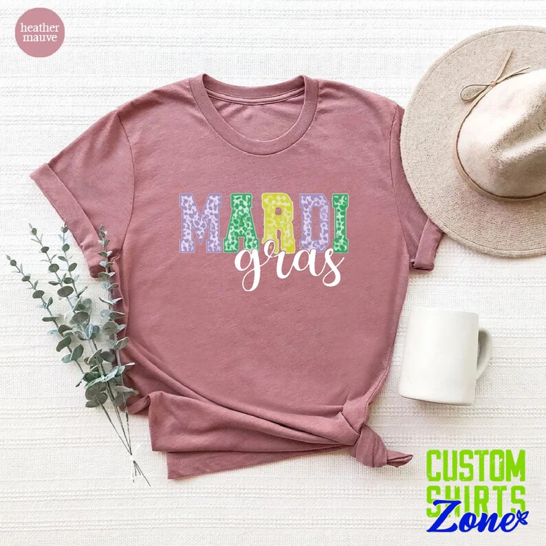 May include: A heather mauve t-shirt with the words "Mardi Gras" in green, purple, and gold letters with a leopard print pattern. The letters are outlined in white.