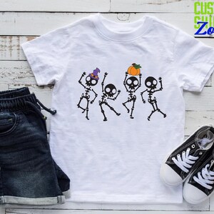 May include: White t-shirt with a graphic of four skeletons dancing. The skeletons are wearing hats and holding a pumpkin. The text "Custom Shirts Zone" is in the top right corner of the image.