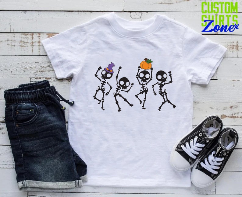 May include: White t-shirt with a graphic of four skeletons dancing. The skeletons are wearing hats and holding a pumpkin. The text "Custom Shirts Zone" is in the top right corner of the image.