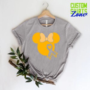 K&ouml;nnte beinhalten: Ein graues T-Shirt mit einer gelben Silhouette von Mickey Mouse mit einer gelben Schleife und einem gelben Band mit einem Herzen in der Mitte. Das Band ist ein Symbol f&uuml;r das Bewusstsein f&uuml;r Kinderkrebs.