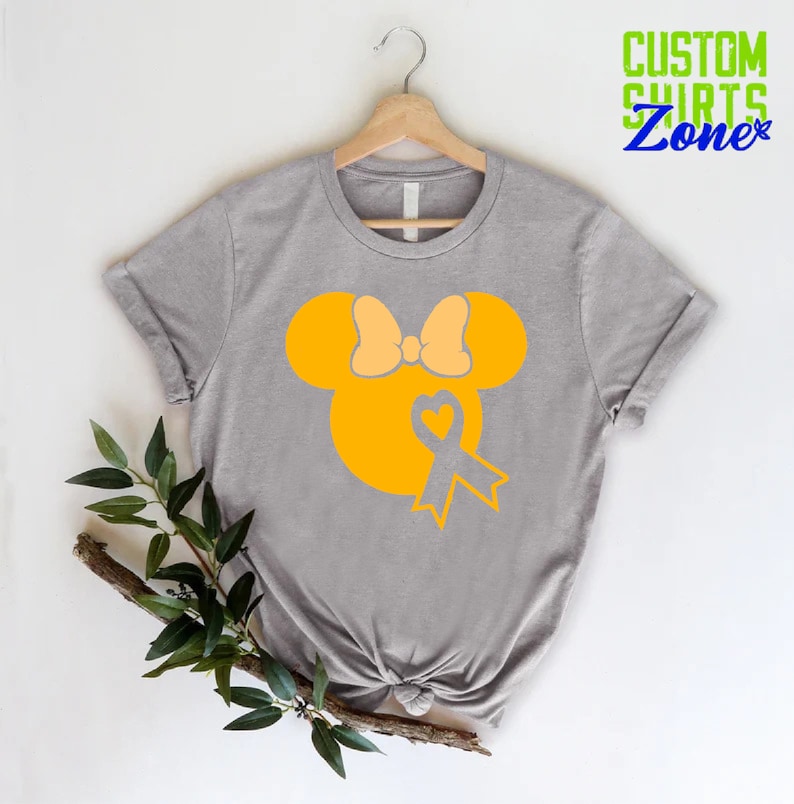 K&ouml;nnte beinhalten: Ein graues T-Shirt mit einer gelben Silhouette von Mickey Mouse mit einer gelben Schleife und einem gelben Band mit einem Herzen in der Mitte. Das Band ist ein Symbol f&uuml;r das Bewusstsein f&uuml;r Kinderkrebs.