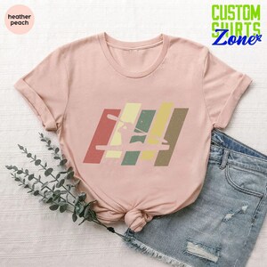 K&ouml;nnte beinhalten: Ein T-Shirt in der Farbe 'Heather Peach' mit einer Retro-Grafik einer Person im Kajak. Die Grafik besteht aus bunten Streifen in den Farben Rot, Gelb, Gr&uuml;n und Braun.