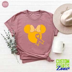 K&ouml;nnte beinhalten: Ein T-Shirt in der Farbe Heide-Mauve mit einer gelben Mickey-Maus-Silhouette, die eine gelbe Schleife und ein gelbes Band mit einem Herzen in der Mitte tr&auml;gt. Das Band ist ein Symbol f&uuml;r das Bewusstsein f&uuml;r Kinderkrebs.