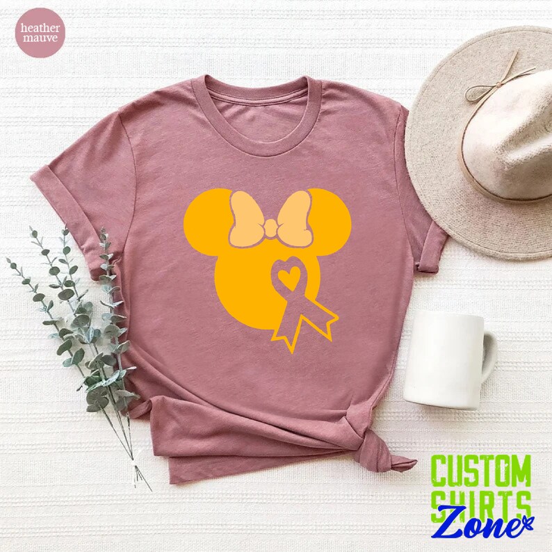 K&ouml;nnte beinhalten: Ein T-Shirt in der Farbe Heide-Mauve mit einer gelben Mickey-Maus-Silhouette, die eine gelbe Schleife und ein gelbes Band mit einem Herzen in der Mitte tr&auml;gt. Das Band ist ein Symbol f&uuml;r das Bewusstsein f&uuml;r Kinderkrebs.