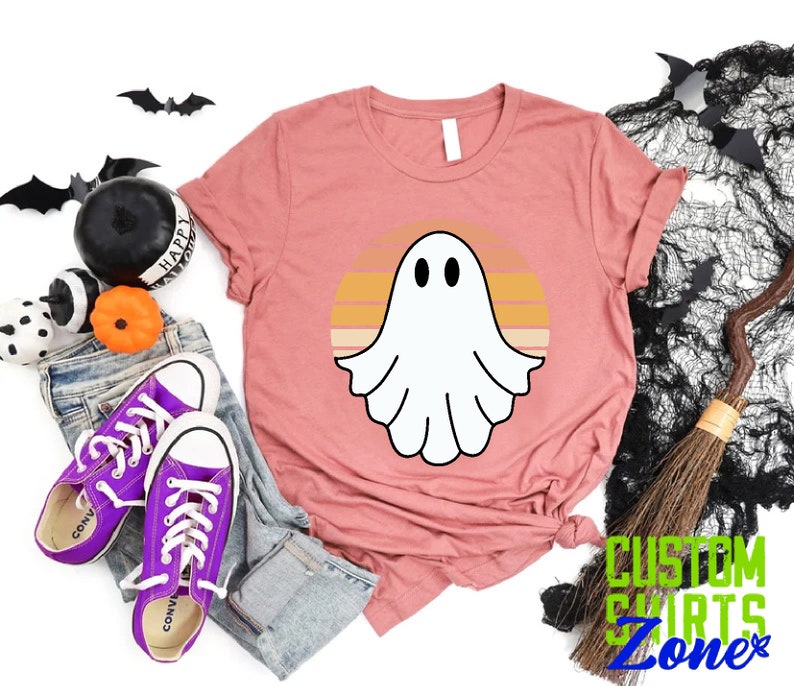 May include: A pink t-shirt with a white ghost silhouette in front of a retro sunset design. The text "CUSTOM SHIRTS ZONE" is visible in the bottom right corner of the image.
