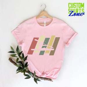 K&ouml;nnte beinhalten: Pinkes T-Shirt mit einer Retro-Grafik einer Person im Kajak. Die Grafik besteht aus bunten Streifen mit einem Used-Look.