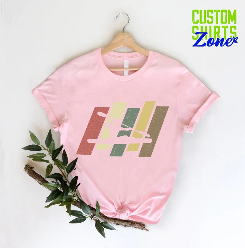 K&ouml;nnte beinhalten: Pinkes T-Shirt mit einer Retro-Grafik einer Person im Kajak. Die Grafik besteht aus bunten Streifen mit einem Used-Look.