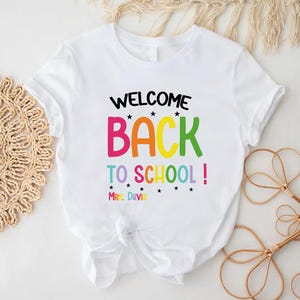 以下が含まれることがあります： カラフルな文字で「WELCOME BACK TO SCHOOL！」と書かれた白いTシャツ。Tシャツには、テキストの下に「Mrs. Davis」という名前も含まれています。Tシャツは下部で結ばれています。