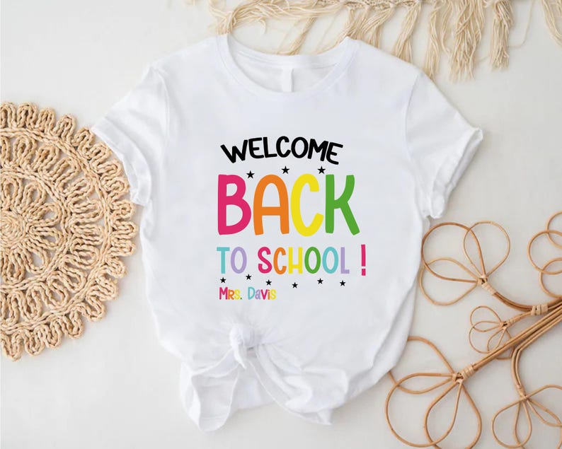 以下が含まれることがあります： カラフルな文字で「WELCOME BACK TO SCHOOL！」と書かれた白いTシャツ。Tシャツには、テキストの下に「Mrs. Davis」という名前も含まれています。Tシャツは下部で結ばれています。