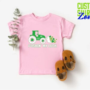 May include: Pink t-shirt with a green tractor and shamrocks, and the text "Pushin' My Luck".