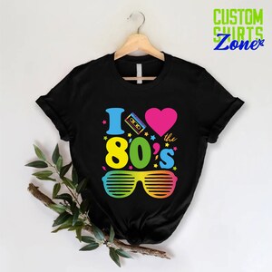 May include: Black t-shirt with a graphic design that says "I love the 80's" with a cassette tape, a heart, and sunglasses. The text is in rainbow colors.