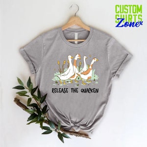 May include: A gray t-shirt with a graphic of four ducks walking in a line. The ducks are white with orange heads and feet. The text "Release the Quacken" is printed below the ducks.