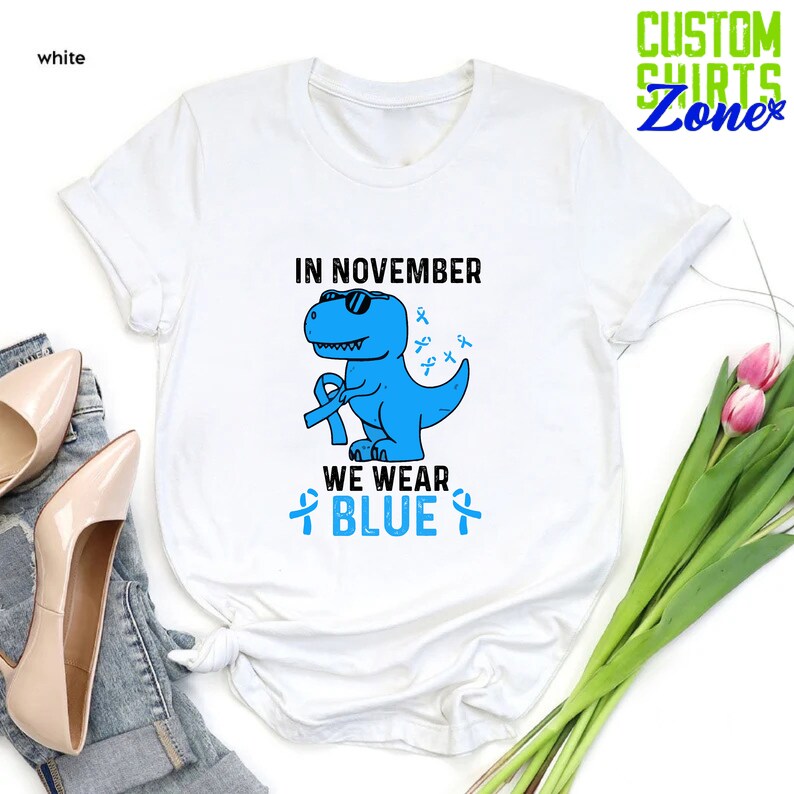May include: White t-shirt with a blue dinosaur wearing sunglasses and holding a blue ribbon. The text "IN NOVEMBER WE WEAR BLUE" is printed on the shirt. The shirt has a crew neck and short sleeves. The words "CUSTOM SHIRTS Zone" are printed in green.