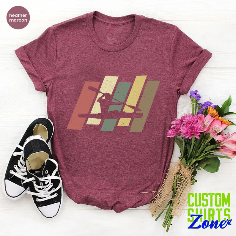 K&ouml;nnte beinhalten: Ein T-Shirt in Heidelbeer-Maroon mit einer Retro-Grafik einer Person im Kajak. Die Grafik besteht aus bunten Streifen in den Farben Gelb, Orange, Gr&uuml;n und Braun.