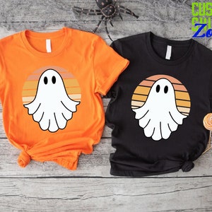 May include: Two t-shirts with a retro sunset design featuring a white ghost. The t-shirts are orange and black.