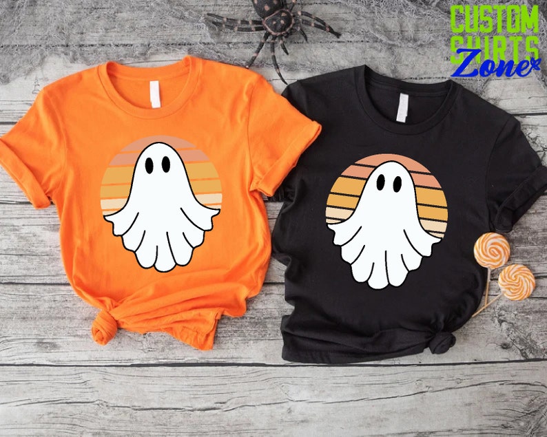 May include: Two t-shirts with a retro sunset design featuring a white ghost. The t-shirts are orange and black.
