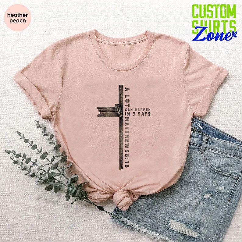 May include: A pink t-shirt with a brown wooden cross graphic. The cross has the text "A LOT CAN HAPPEN IN 3 DAYS MATTHEW 28:16" on it.