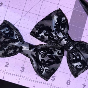 May include: Two black and silver bow ties with a damask pattern. The bows are made of a shiny fabric and are laying on a purple cutting mat. The bows are a classic accessory.