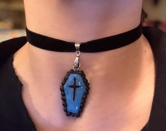 May include: A black velvet choker necklace with a coffin pendant. The pendant is light blue with a black cross and is framed by black beads. The necklace is a gothic accessory.