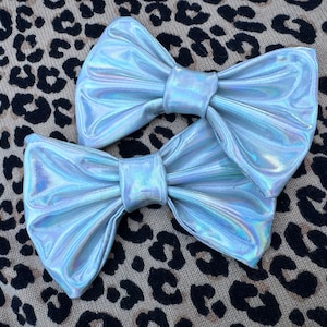 May include: Two iridescent silver bow hair clips. The bows are made of a shiny, reflective fabric and are arranged in a classic bow shape.