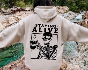 Może przedstawiać: Beżowa bluza z kapturem z czarnym nadrukiem szkieletu trzymającego kubek z kawą z napisem "Coffee". Tekst "Staying Alive" jest nadrukowany nad grafiką.