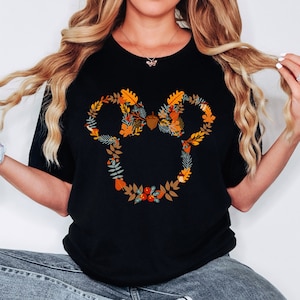 May include: Black t-shirt with a floral Mickey Mouse design. The design features autumn leaves in orange, yellow, and green, with a brown acorn at the centre. The shirt has a crew neck.