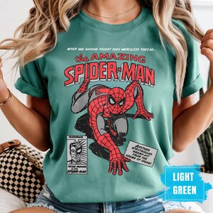 May include: Light green t-shirt featuring a vintage comic book design of Spider-Man. The graphic includes the text "the AMAZING SPIDER-MAN" and "PETER PARKER alias SPIDER-MAN". The shirt also has the text "NEVER HAS ANYONE FOUGHT SUCH MERCILESS FOES AS..." and "ANOTHER SPECTACULAR SMASH-HIT FROM THE HOUSE OF IDEAS!"