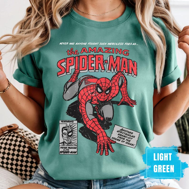 May include: Light green t-shirt featuring a vintage comic book design of Spider-Man. The graphic includes the text "the AMAZING SPIDER-MAN" and "PETER PARKER alias SPIDER-MAN". The shirt also has the text "NEVER HAS ANYONE FOUGHT SUCH MERCILESS FOES AS..." and "ANOTHER SPECTACULAR SMASH-HIT FROM THE HOUSE OF IDEAS!"