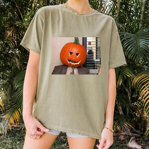 May include: A green t-shirt with a graphic of a person wearing a jack-o'-lantern on their head. The person is wearing a suit and tie. The graphic is in a square frame.