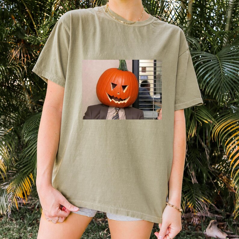 May include: A green t-shirt with a graphic of a person wearing a jack-o'-lantern on their head. The person is wearing a suit and tie. The graphic is in a square frame.