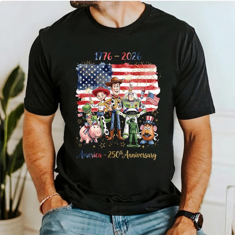 Op de afbeelding: Zwart T-shirt met een kleurrijke afbeelding van animatiefiguren voor een Amerikaans vlagontwerp. De tekst "1776-2026" en "America - 250th Anniversary" staan ook op het shirt.