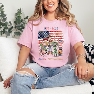Op de afbeelding: Lichtroze T-shirt met een afbeelding van de Amerikaanse vlag en Toy Story-personages. Het shirt toont de tekst "1776-2026" en "America - 250th Anniversary" in goud. Het T-shirt heeft een ronde hals en is gemaakt van een zacht materiaal.