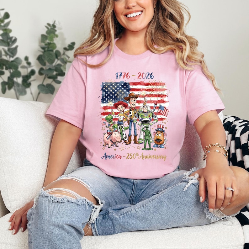 Op de afbeelding: Lichtroze T-shirt met een afbeelding van de Amerikaanse vlag en Toy Story-personages. Het shirt toont de tekst "1776-2026" en "America - 250th Anniversary" in goud. Het T-shirt heeft een ronde hals en is gemaakt van een zacht materiaal.
