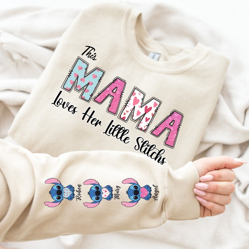 May include: A beige sweatshirt with the text "This Mama Loves Her Little Stitches" in pink and blue letters with hearts. The sleeve has three blue Stitch characters with hearts and the names "Robin", "Mary", and "Angel" underneath.
