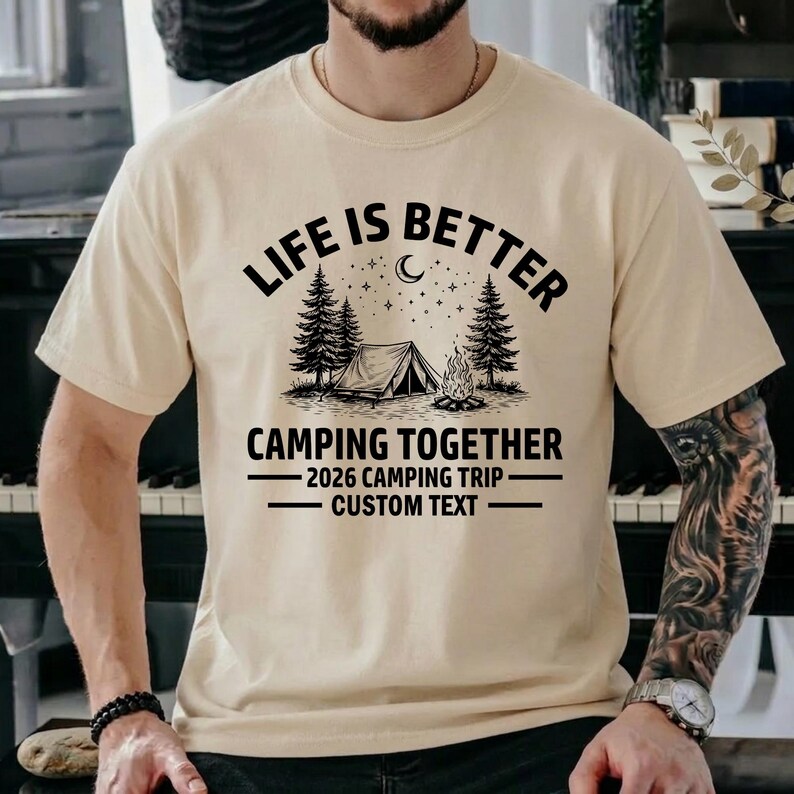 Puede incluir: Una camiseta beige con un dise&ntilde;o tem&aacute;tico de camping. El dise&ntilde;o incluye el texto "LIFE IS BETTER CAMPING TOGETHER" encima de una tienda de campa&ntilde;a, &aacute;rboles y una fogata. Debajo, se lee "2026 CAMPING TRIP CUSTOM TEXT".