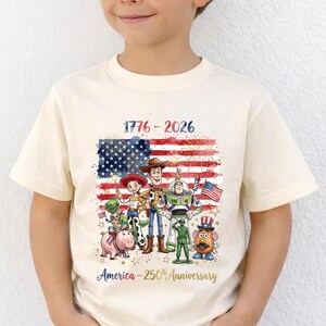 Op de afbeelding: Cr&egrave;mekleurig T-shirt met een patriottisch ontwerp. Het heeft de tekst "1776-2026" en "America - 250th Anniversary". De afbeelding bevat de Amerikaanse vlag en stripfiguren. Het shirt wordt door een kind gedragen.