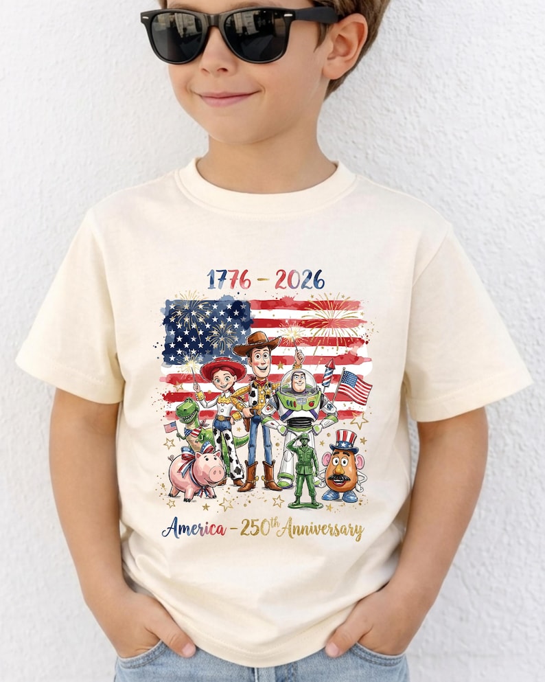 Op de afbeelding: Cr&egrave;mekleurig T-shirt met een patriottisch ontwerp. Het heeft de tekst "1776-2026" en "America - 250th Anniversary". De afbeelding bevat de Amerikaanse vlag en stripfiguren. Het shirt wordt door een kind gedragen.