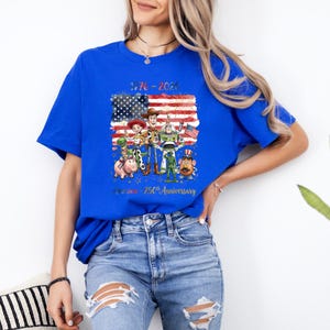Op de afbeelding: Koningsblauw T-shirt met een afbeelding van tekenfilmfiguren voor een Amerikaanse vlag. De tekst "1776-2026" en "America - 250th Anniversary" staan op het shirt. Het shirt is gemaakt van een zacht materiaal.
