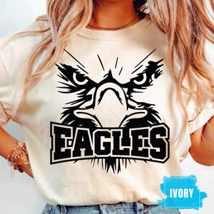 May include: An ivory t-shirt featuring a bold black graphic of an eagle's face with sharp, angular lines. Below the eagle, the word "EAGLES" is printed in a large, blocky, black font. The design is centered on the front of the shirt.