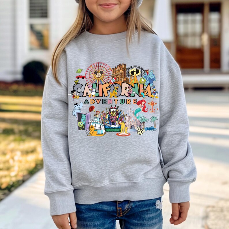 May include: A light gray sweatshirt featuring a colorful graphic that reads "CALIFORNIA ADVENTURE" with various cartoon characters and amusement park imagery. The design includes a Ferris wheel, castle, and other theme park elements.