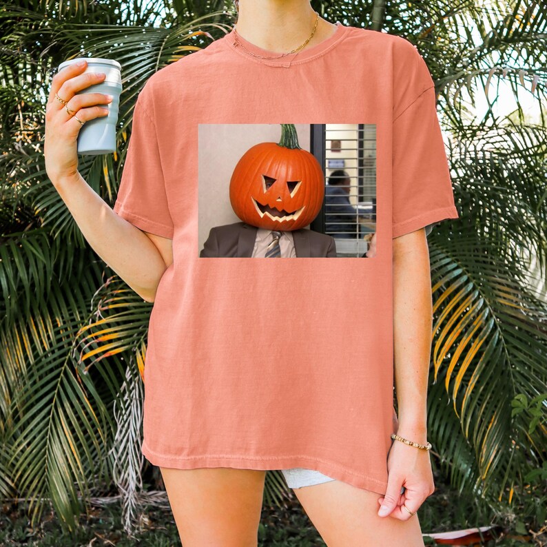 May include: A coral colored t-shirt with a graphic of a person with a pumpkin for a head. The pumpkin has a carved face and is wearing a tie.