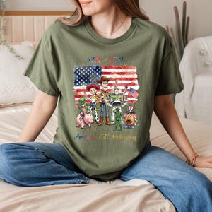 Op de afbeelding: Olijfgroen t-shirt met een afbeelding van de Amerikaanse vlag, vuurwerk en cartoonfiguren. De tekst "1776-2026" en "America 250th Anniversary" zijn ook zichtbaar. Het t-shirt wordt door een persoon gedragen.