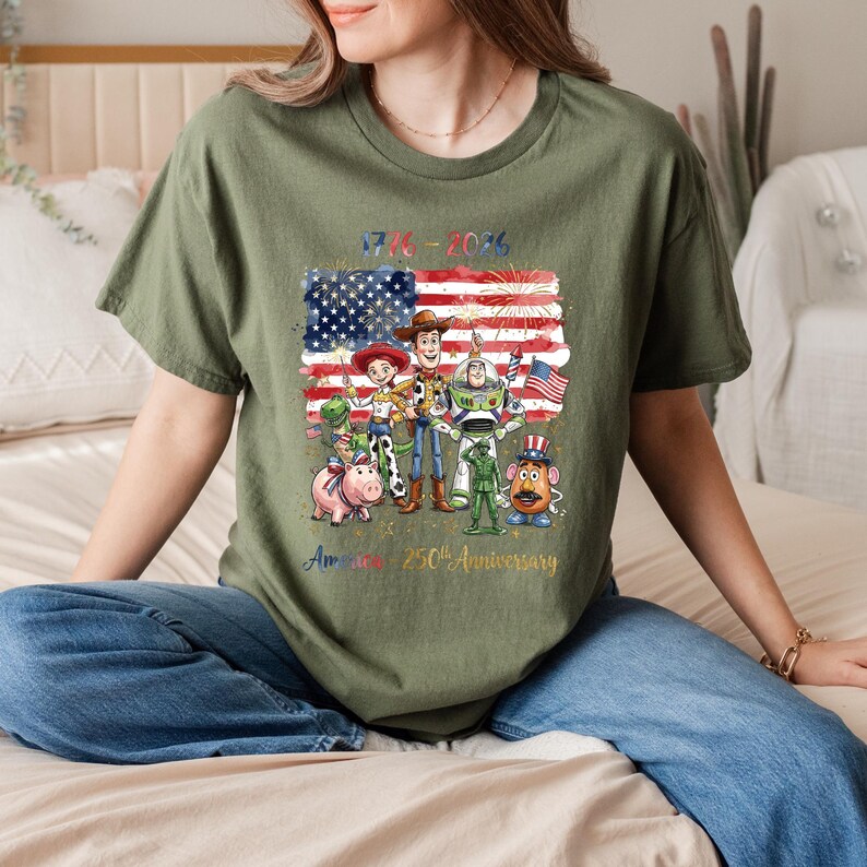 Op de afbeelding: Olijfgroen t-shirt met een afbeelding van de Amerikaanse vlag, vuurwerk en cartoonfiguren. De tekst "1776-2026" en "America 250th Anniversary" zijn ook zichtbaar. Het t-shirt wordt door een persoon gedragen.