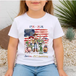 Op de afbeelding: Wit T-shirt met een kleurrijke afbeelding van de Amerikaanse vlag en Toy Story-personages. De tekst "1776-2026" en "America - 250th Anniversary" zijn ook opgenomen.
