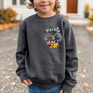 K&ouml;nnte beinhalten: Ein dunkelgrauer Pullover mit einem wei&szlig;en Aufdruck von Mickey Mouse und seinen Freunden mit Feuerwerk. Der Text "KELVIN" steht &uuml;ber dem Aufdruck.