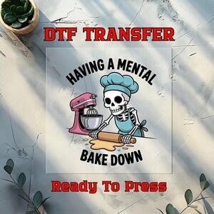 May include: DTF transfer featuring a skeleton baker with a chef's hat and apron, rolling dough. A pink stand mixer is to the left. Text reads "HAVING A MENTAL BAKE DOWN" and "READY TO PRESS" in red.