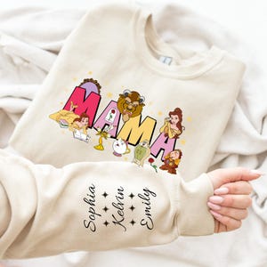 May include: A beige sweatshirt featuring a graphic of the word "MAMA" in colourful letters with characters from the Disney film Beauty and the Beast. The sleeve has the names "Sophia", "Kelvin", and "Emily" written in black.