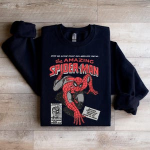 May include: Black sweatshirt with a vintage comic book design featuring Spider-Man. The text reads "NEVER HAS ANYONE FOUGHT SUCH MERCILESS FOES AS... the AMAZING SPIDER-MAN".